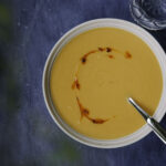 Bataattikeitto on helppo ja herkullinen arkiruoka. Kuvassa oranssi keitto on vaalealla lautasella. Keiton päällä on chiliöljyä. Kuvan tausta on sininen.