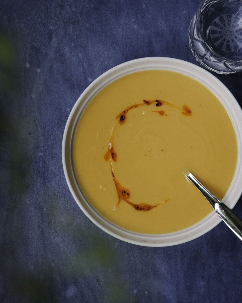 Bataattikeitto on helppo ja herkullinen arkiruoka. Kuvassa oranssi keitto on vaalealla lautasella. Keiton päällä on chiliöljyä. Kuvan tausta on sininen.