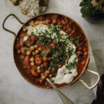 Kikherneet harissa-tomaattikastikkeessa on helppo arkiruoka tai lisäke. Kuvassa ruoka on pyöreässä metallivuoassa. Päällä on maustamatonta jugua, yrittisilppua ja oliiviöljyä.