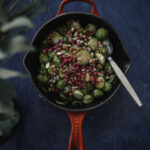 Paahdetut ruusukaalit vaahterasiirapilla on helppo ja herkullinen lisäke. Kuvassa paistos on pienellä paistinpannulla, jonka ulkopuoli on kirkkaan punainen. Ruusukaalien päällä on mantelirouhetta ja granaattiomenansiemeniä. Kuva on otettu ylhäältäpäin ja kuvan tausta on tummansininen.