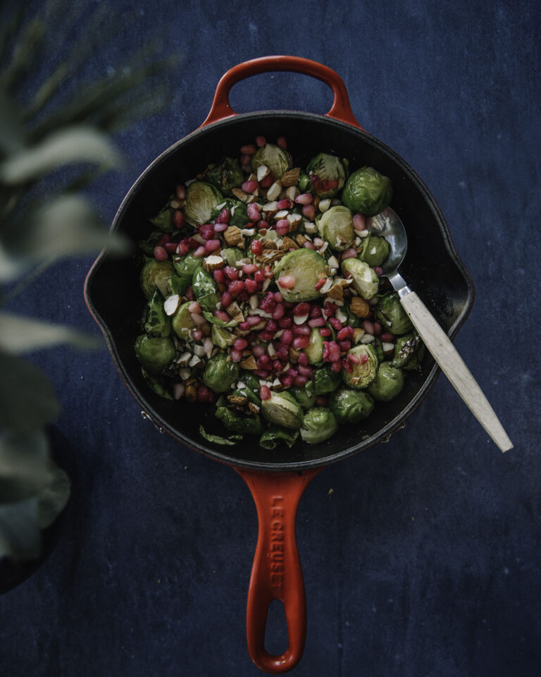 Paahdetut ruusukaalit vaahterasiirapilla on helppo ja herkullinen lisäke. Kuvassa paistos on pienellä paistinpannulla, jonka ulkopuoli on kirkkaan punainen. Ruusukaalien päällä on mantelirouhetta ja granaattiomenansiemeniä. Kuva on otettu ylhäältäpäin ja kuvan tausta on tummansininen.