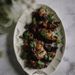 Gochujang-kana on ihanan helppo ruoka, joka valmistuu grillissä tai uunissa. Kuvassa ruoka on soikealla lautasella. Päällä on korianterin lehtiä. Vasemmassa yläkulmassa on epäselvänä niittykukkakimppu. Kuva on otettu ylhäältäpäin.
